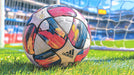 Close-up of a soccer ball with abstract red, blue, and yellow brushstroke patterns sitting on a bright green grass field.