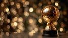 A close-up of a gleaming gold soccer ball trophy against a blurred, golden bokeh light background.
