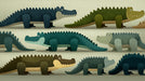 A whimsical pattern of stylized crocodiles in deep navy, olive, and ochre on a textured sand background.