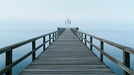 A long wooden pier stretching into a foggy, misty pale blue sea and sky landscape.