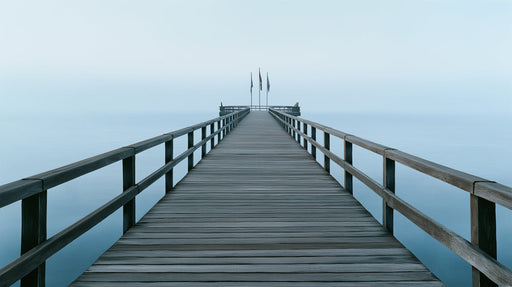 A long wooden pier stretching into a foggy, misty pale blue sea and sky landscape.