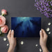 Person holding a card with underwater scene of sharks and people, surrounded by flowers and petals on a dark surface.