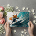 Person holding a card with a cartoon fox surfing on a wave, surrounded by flowers and petals.